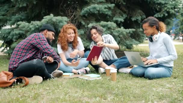 Estudantes alegres estudando ao ar livre com livros e laptop conversando na grama — Vídeo de Stock