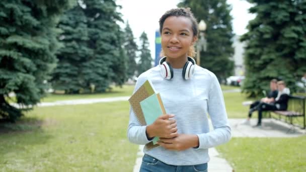 Linda menina afro-americana andando ao ar livre no parque segurando livros sorrindo — Vídeo de Stock