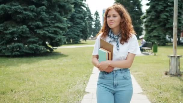 Pretty redhead bayan öğrenci sonbaharda gülümseyerek kitap tutan parkta yürüyüş — Stok video