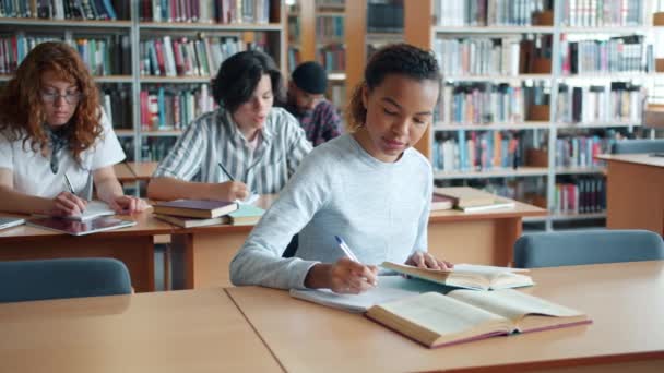 Αργή κίνηση του αρκετά μικτή φυλή κορίτσι μελέτη στη βιβλιοθήκη με την ομάδα των ανθρώπων — Αρχείο Βίντεο