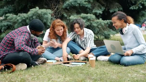 Захоплені студенти читають книги і використовують ноутбук у парку сміються розваги — стокове відео