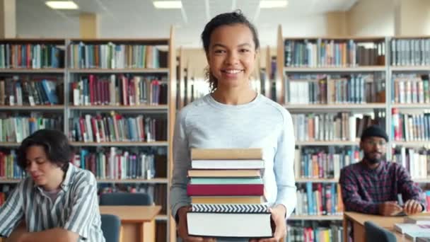 Lycklig afrikansk amerikansk kvinna går i skolans bibliotek håller böcker leende — Stockvideo