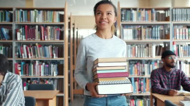 Довольно смешанной расы студентка несет книги ходить в библиотеке улыбаясь — стоковое видео