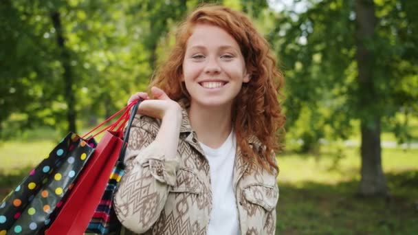 Movimento lento de menina alegre com sacos de compras em pé no parque sozinho sorrindo — Vídeo de Stock