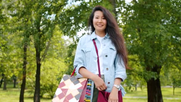 Retrato de chica asiática bonita de pie en el parque con bolsas de compras sonriendo — Vídeos de Stock