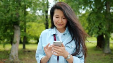 Şehir parkında akıllı telefon kullanan çekici Asyalı kadın ekrana dokunuyor gülümseyerek