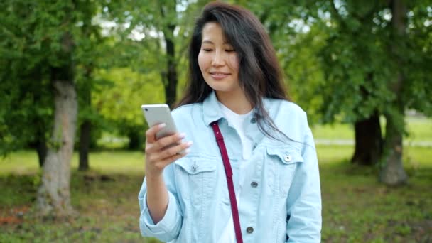 Šťastná asijská žena, mluvící na mobilním telefonu a usmívající se venku na městském parku — Stock video