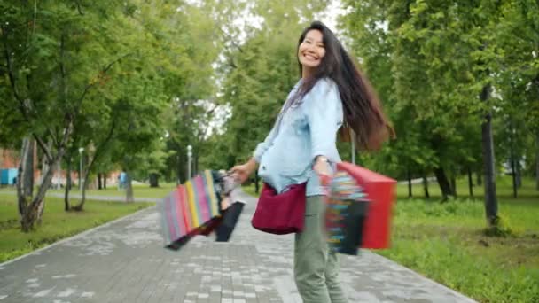 Retrato de chica asiática feliz girando en parque sosteniendo bolsas de compras divirtiéndose — Vídeos de Stock