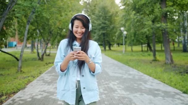 Счастливая азиатка в наушниках танцует на улице в парке со смартфоном — стоковое видео