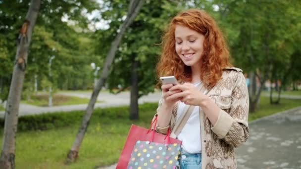 Mouvement lent de l'adolescent marchant dans le parc avec des sacs à provisions à l'aide d'un smartphone — Video
