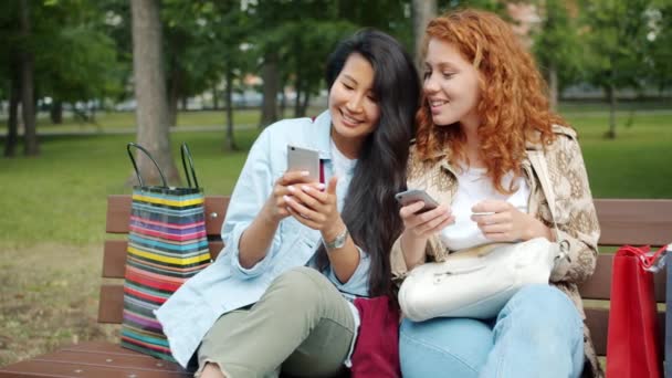 Веселые девушки с помощью смартфонов болтают расслабляясь на скамейке в парке вместе — стоковое видео