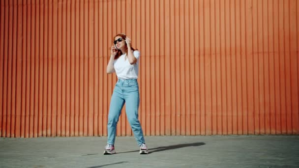 Chica alegre en auriculares y gafas de sol bailando al aire libre en la calle divirtiéndose — Vídeo de stock
