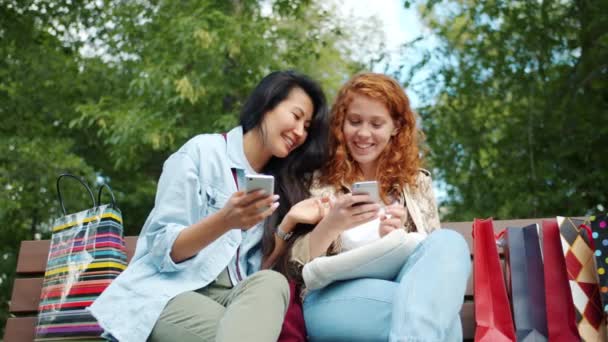 Радостные друзья, пользующиеся смартфонами на открытом воздухе в парке смеются над обсуждением контента — стоковое видео