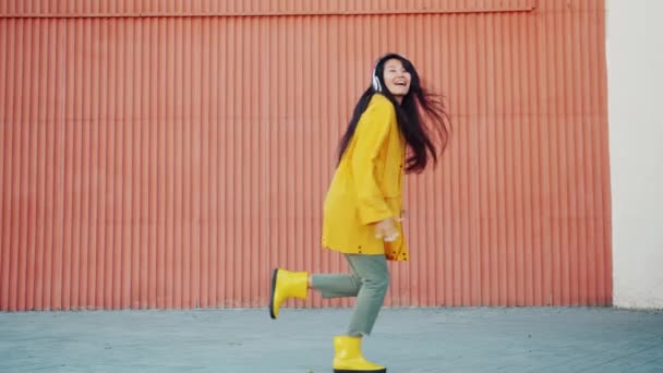 Vidám ázsiai hölgy esőkabát és gumi csizma táncoló szabadban szórakozik — Stock videók