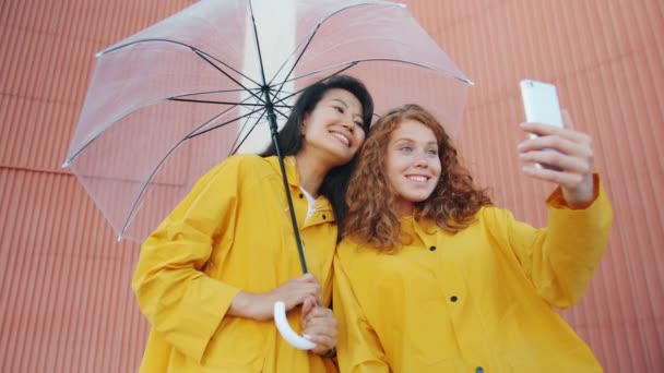 Glückliche Freundinnen in Regenmänteln mit Regenschirm für Selfies im Freien — Stockvideo