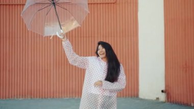 Yağmurluk dans mutlu Asyalı kız kamera bakarak şemsiye tutarak açık havada dans