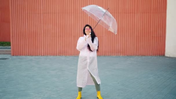 Медленное движение азиатки в плаще и резиновых сапогах, танцующей с зонтиком — стоковое видео