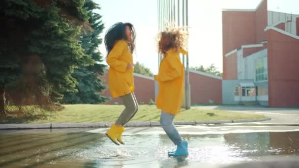 Adolescentes felizes em capas de chuva e gomas pulando na poça se divertindo rindo — Vídeo de Stock