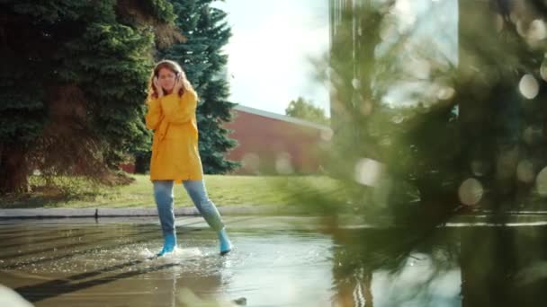 Student in raincoat, gumboots jumping in puddle listening to music in headphones — Stock Video