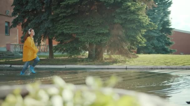 Happy teen in raincoat and gumboots running in puddles wearing headphone — Stok Video