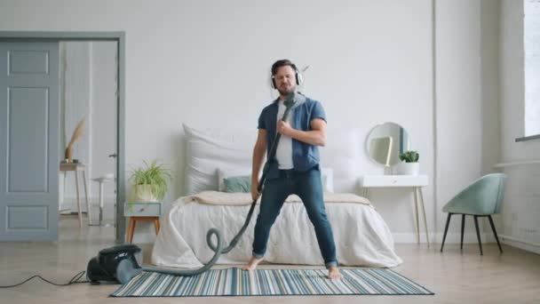 Feliz joven en auriculares cantando en aspiradora bailando limpiando casa — Vídeo de stock