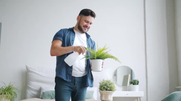 Красивый парень поливает растения дома, используя спрей бутылку наслаждаясь домашней работой — стоковое видео