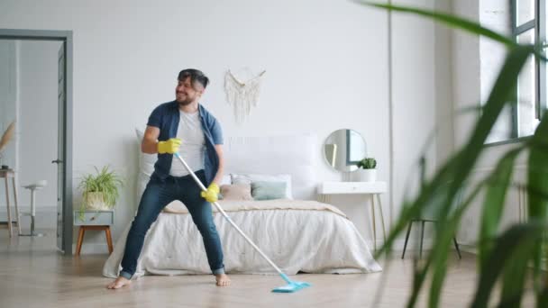 Веселий молодий чоловік танцює співає миття підлоги в квартирі з пластиковим мопедом — стокове відео