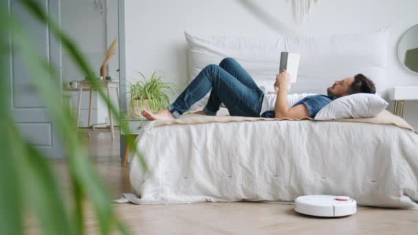 Hombre despreocupado leyendo libro en la cama mientras aspiradora robótica piso de aspiración — Vídeos de Stock