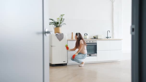 Красивая девушка в наушниках танцует на кухне, выполняя домашнюю работу — стоковое видео