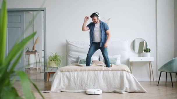 Hombre en auriculares bailando en la cama mientras aspiradora robótica piso de aspiración — Vídeos de Stock