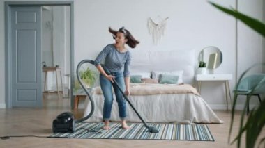 Neşeli genç kız yatak odasında elektrikli süpürge kullanarak şarkı söyleyip dans ediyor.