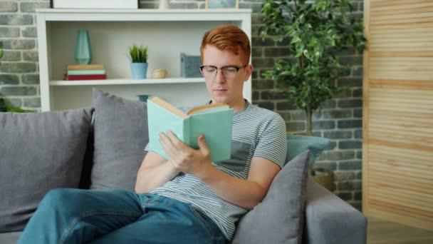 Beau gars dans des lunettes livre de lecture et souriant assis sur le canapé dans l'appartement — Video