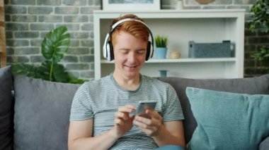 Neşeli adam kulaklıkla müzik dinliyor ve evde akıllı telefon kullanıyor.