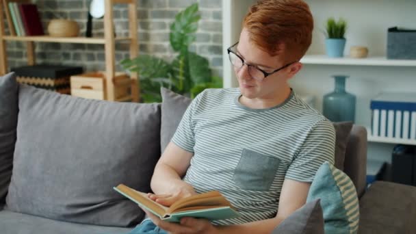 Glimlachende jongeman in bril leesboek zittend op de bank in appartement — Stockvideo