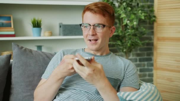 Счастливый молодой человек записывает голосовое сообщение, улыбаясь, используя смартфон дома — стоковое видео