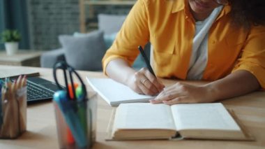 Masada kitap okuyan bir kadın not defterine el yazısıyla yazarken yakın plan.