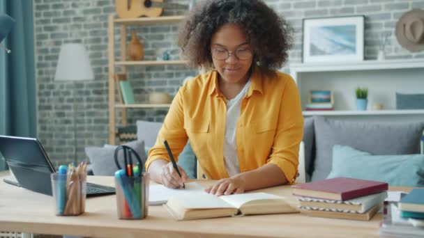 Улыбающаяся молодая женщина пишет в блокноте на столе дома, работая над проектом — стоковое видео