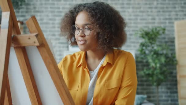 Cámara lenta de la imagen de la pintura de la muchacha afroamericana en casa trabajando con caballete — Vídeos de Stock
