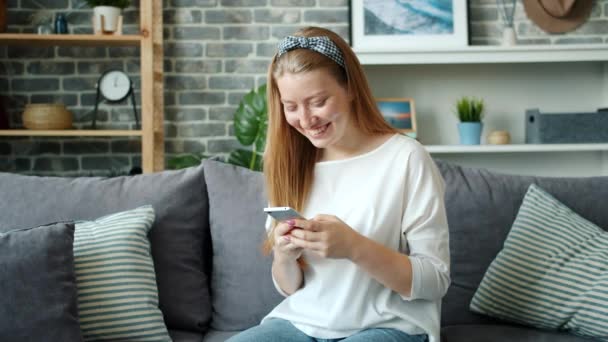 Hermosa chica rubia usando teléfono inteligente y riendo relajarse en el sofá en casa — Vídeo de stock