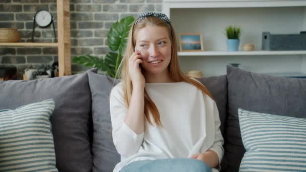 Радісна дівчина спілкується на мобільному телефоні посміхаючись сидячи на дивані в сучасній квартирі — стокове відео