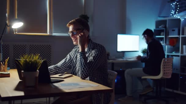 Senyum manajer muda berbicara di telepon genggam di tempat kerja di kantor gelap — Stok Video