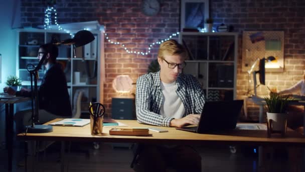 Счастливый парень использует ноутбук в темном офисе, наслаждаясь успехом в бизнесе, улыбаясь — стоковое видео