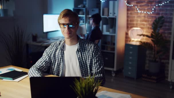 Movimento lento do homem de óculos olhando para a câmera e sorrindo no escritório à noite — Vídeo de Stock