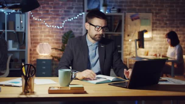 Стресс парень с помощью ноутбука, а затем трогать голову чувство головной боли в темном офисе — стоковое видео