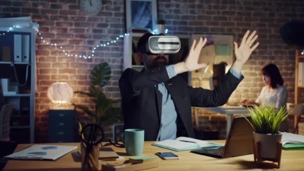 Hombre usando gafas de realidad virtual moviendo brazos sentado en el escritorio en la oficina oscura — Vídeos de Stock