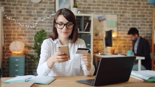 Радостная женщина делает покупки онлайн с помощью кредитной карты с помощью смартфона в офисе — стоковое видео