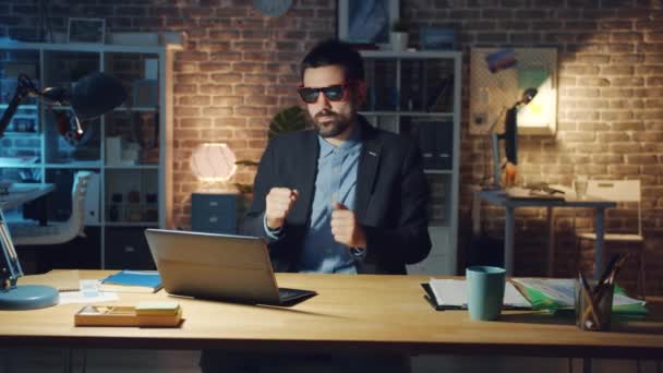 Fröhlicher Mann mit Brille genießt Musik bei der Arbeit mit Laptop tanzen im dunklen Büro — Stockvideo