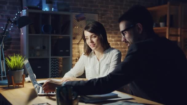 Cheerful young people discussing work pointing at laptop screen in dark office — ストック動画