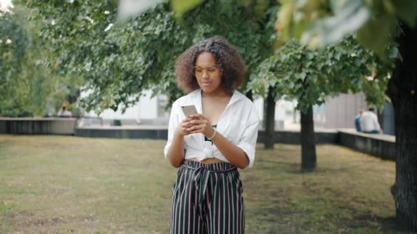 Portrét veselé afroamerické dívky pomocí smartphone venku v městském parku — Stock video