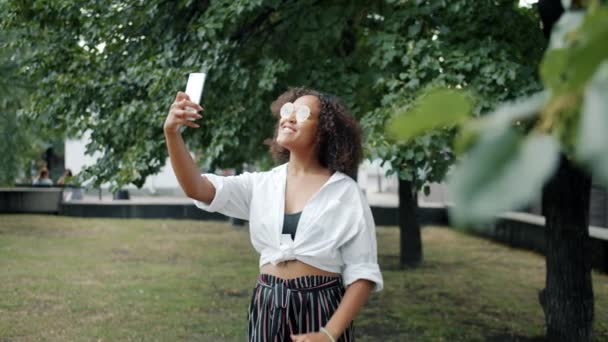 Женщина смешанной расы делает селфи в городском парке, держа камеру смартфона в позе — стоковое видео